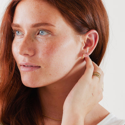 A close-up of a woman's face showcasing a gold hoop earring and two gold square bezel-set gemstone studs, one with a warm red stone and one with an amber stone, paired with matching rings.