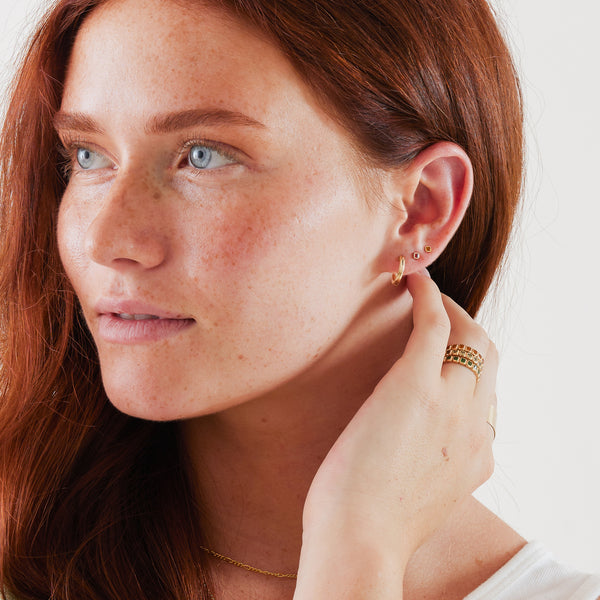 A close-up of a woman's face showcasing a gold hoop earring and two gold square bezel-set gemstone studs, one with a warm red stone and one with an amber stone, paired with matching rings.