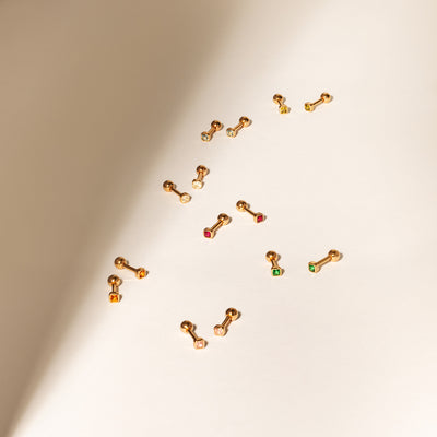 Multiple pairs of gold stud earrings laying on a solid beige surface, each with different colors of square bezel-set gemstones.
