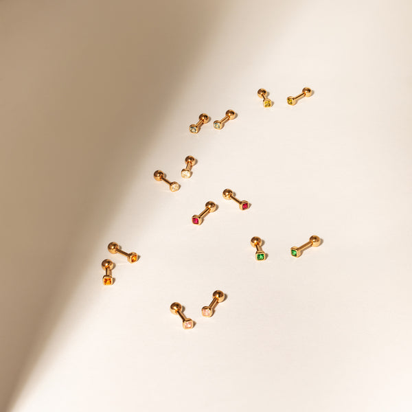 Multiple pairs of gold stud earrings laying on a solid beige surface, each with different colors of square bezel-set gemstones.