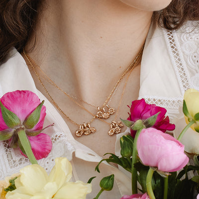 A woman in a white lace collar top holding pink and yellow flowers, wearing three gold pendant necklaces with each having three small flower charms.