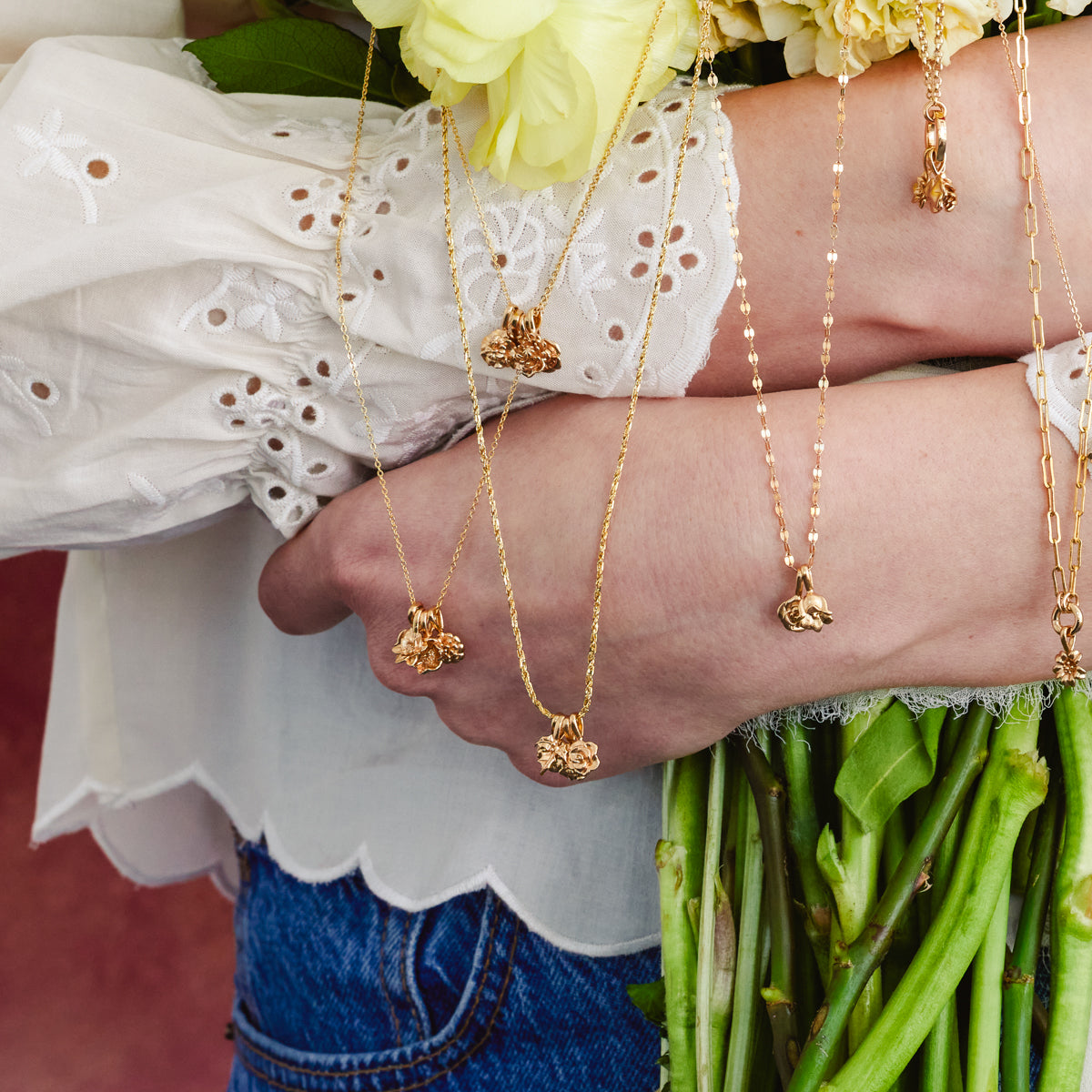 A woman's arms holding a bouquet of flowers, with multiple different gold chain necklaces with small flower charms hanging over her arms.
