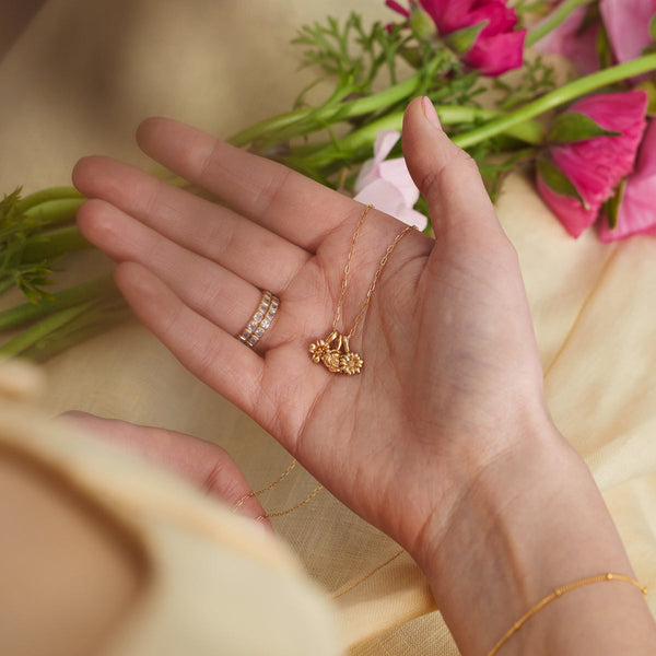 A woman's hand in her lap, with pink flowers blurred in the background, holding a gold pendant necklace with three small flower charms.