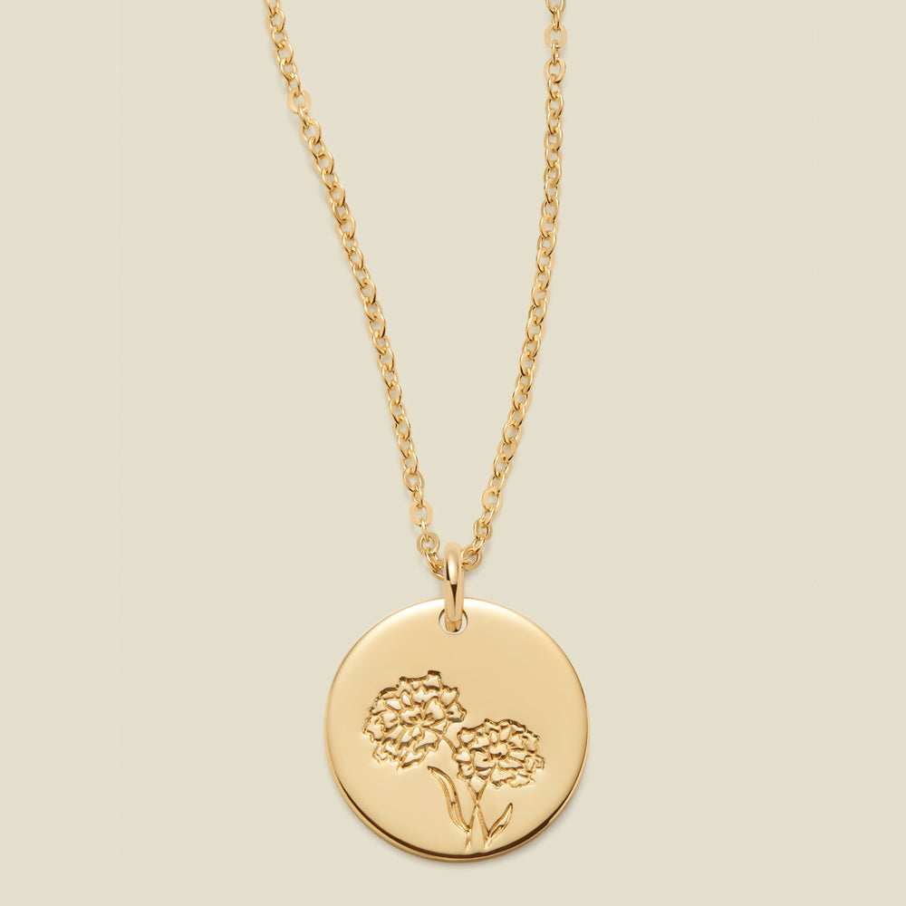 A gold chain necklace with a round gold disc pendant featuring an engraved marigold flower design, displayed against a solid light beige background.