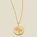 A gold chain necklace with a round gold disc pendant featuring an engraved marigold flower design, displayed against a solid light beige background.