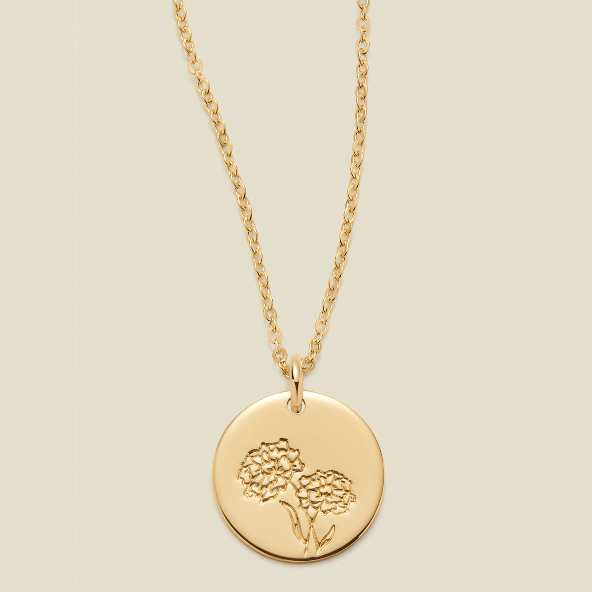 A gold chain necklace with a round gold disc pendant featuring an engraved marigold flower design, displayed against a solid light beige background.