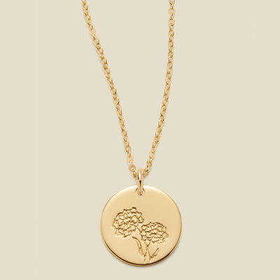 A gold chain necklace with a round gold disc pendant featuring an engraved marigold flower design, displayed against a solid light beige background.