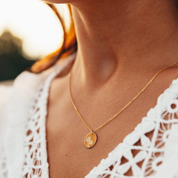 A close-up of a person in a white crotchet top wearing a delicate gold pendant chain necklace with a round flat disc, engraved with a marigold flower design.
