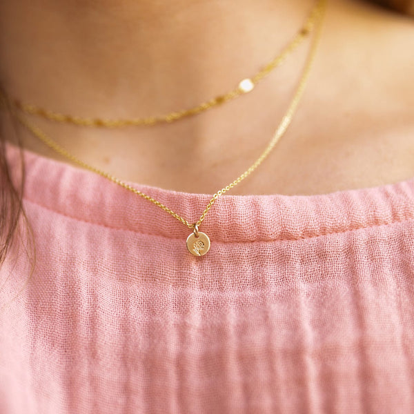 A close-up of a person in a pink linen top wearing a gold pendant necklace with a small round flat disc charm. The disc has a marigold flower engraving.