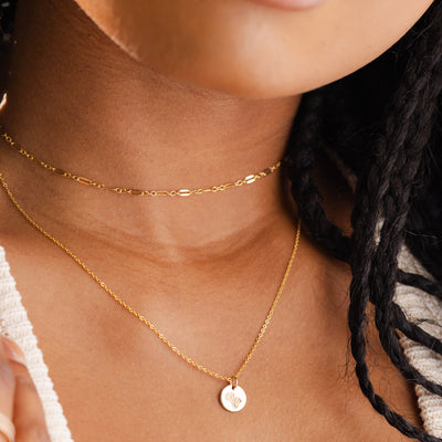 A close-up of a person wearing a thin textured gold choker layered with a delicate gold pendant chain necklace featuring a round flat floral engraved disc.