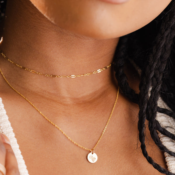 A close-up of a person wearing a thin textured gold choker layered with a delicate gold pendant chain necklace featuring a round flat floral engraved disc.