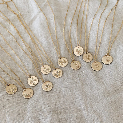 Twelve gold pendant chain necklaces, each with a round flat disc engraved with various floral motifs, laying on a light beige linen background.