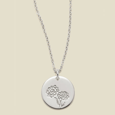 A silver chain necklace with a round silver disc pendant featuring an engraved marigold flower design, displayed against a solid light beige background.