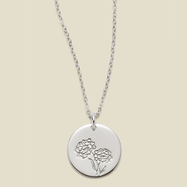 A silver chain necklace with a round silver disc pendant featuring an engraved marigold flower design, displayed against a solid light beige background.