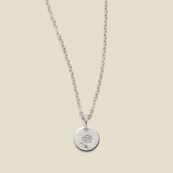 A silver chain necklace with a round silver disc pendant featuring an engraved marigold flower design, displayed against a solid light beige background.