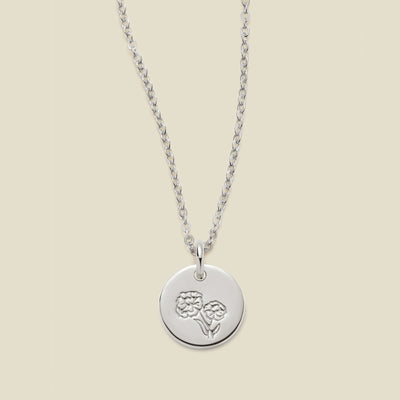 A silver chain necklace with a round silver disc pendant featuring an engraved marigold flower design, displayed against a solid light beige background.
