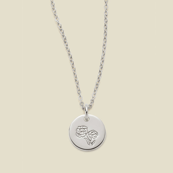 A silver chain necklace with a round silver disc pendant featuring an engraved marigold flower design, displayed against a solid light beige background.