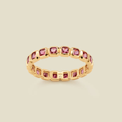 A gold eternity ring featuring a continuous row of square cut pink gemstones, shown on a solid light beige background.