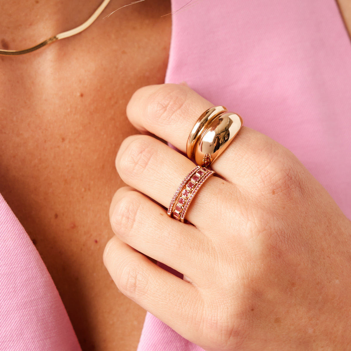 A close-up of a person in a pink shirt resting their hand on their chest, showcasing two polished gold bubble rings and multiple stacked gold rings with different sizes and shapes of pink gemstones.