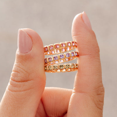 A hand holding three gold eternity rings between two fingers, each ring with either pink, light purple, or light green square cut gemstones in a single continuous row.