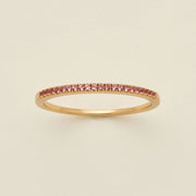 A gold ring featuring a single row of small round-cut pink gemstones, photographed on a clean, solid, warm off-white background.