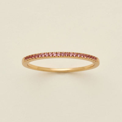A gold ring featuring a single row of small round-cut pink gemstones, photographed on a clean, solid, warm off-white background.