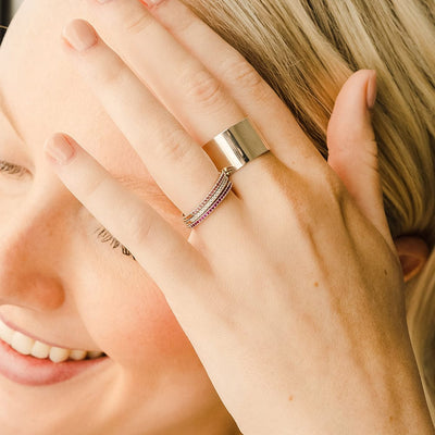 A close-up of a hand resting on someone's face, showcasing a thick polished gold cigar band ring and three stacked thin gold rings, each with a single row of small gemstones in the colors pink, white, and red.