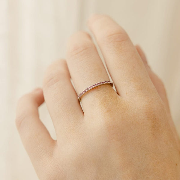 A close-up of a hand wearing a thin gold band ring with a single row of small pink gemstones, set against a light beige background.