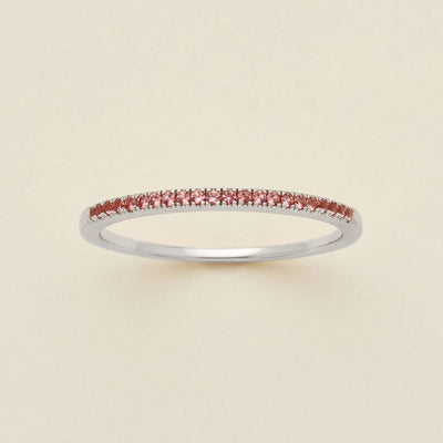 A silver ring featuring a single row of small round-cut pink gemstones, photographed on a clean, solid, warm off-white background.