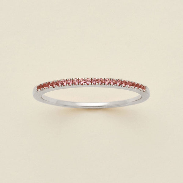 A silver ring featuring a single row of small round-cut pink gemstones, photographed on a clean, solid, warm off-white background.