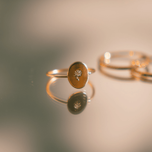 A delicate gold wire ring with a flat oval in the center, engraved with a flower and sitting on a mirror surface with other rings blurred in the background.
