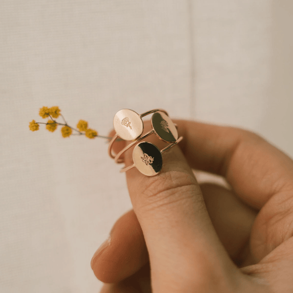 A person's thumb holding three gold wire rings with flat ovals, each engraved with a different floral motif with a light beige background and a small sprig of yellow flowers.