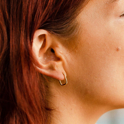 A close-up of a person's ear wearing a gold rounded oval hoop earring.