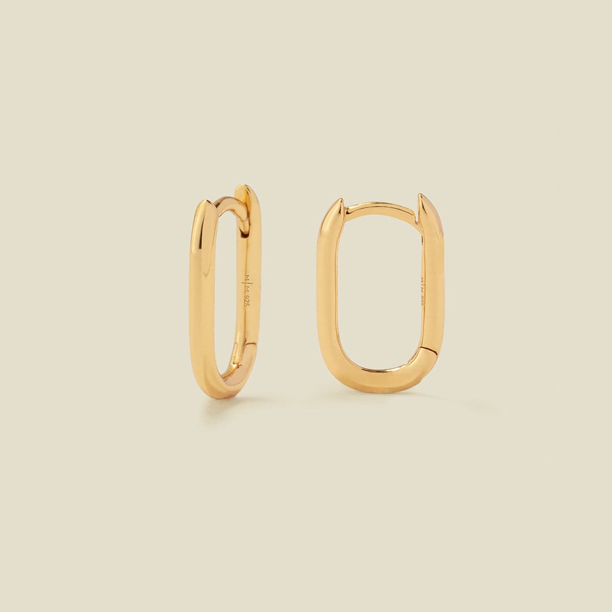 A pair of gold oval hoops with a hinge closure, shown on a solid light beige background.