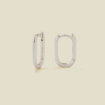 A pair of silver oval hoops with a hinge closure, shown on a solid light beige background.