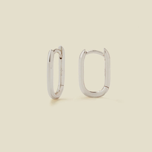 A pair of silver oval hoops with a hinge closure, shown on a solid light beige background.