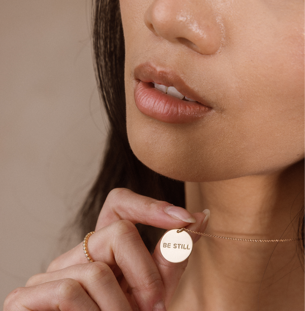 Close-up of a woman wearing a delicate gold chain necklace with a round gold disc pendant engraved with the words 'BE STILL'.