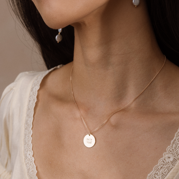 A close-up lifestyle shot of a model wearing a delicate gold chain necklace with a round engraved disc pendant that reads 'FAITH / FEAR'. The necklace is shown on the model's neck, paired with a cream lace-trimmed top.