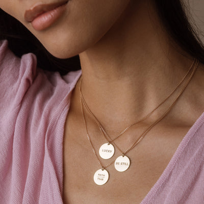 Close-up of a person wearing layered gold necklaces with circular gold-toned pendants engraved with 'LUCKY', 'BE STILL', and 'FAITH / FEAR'.