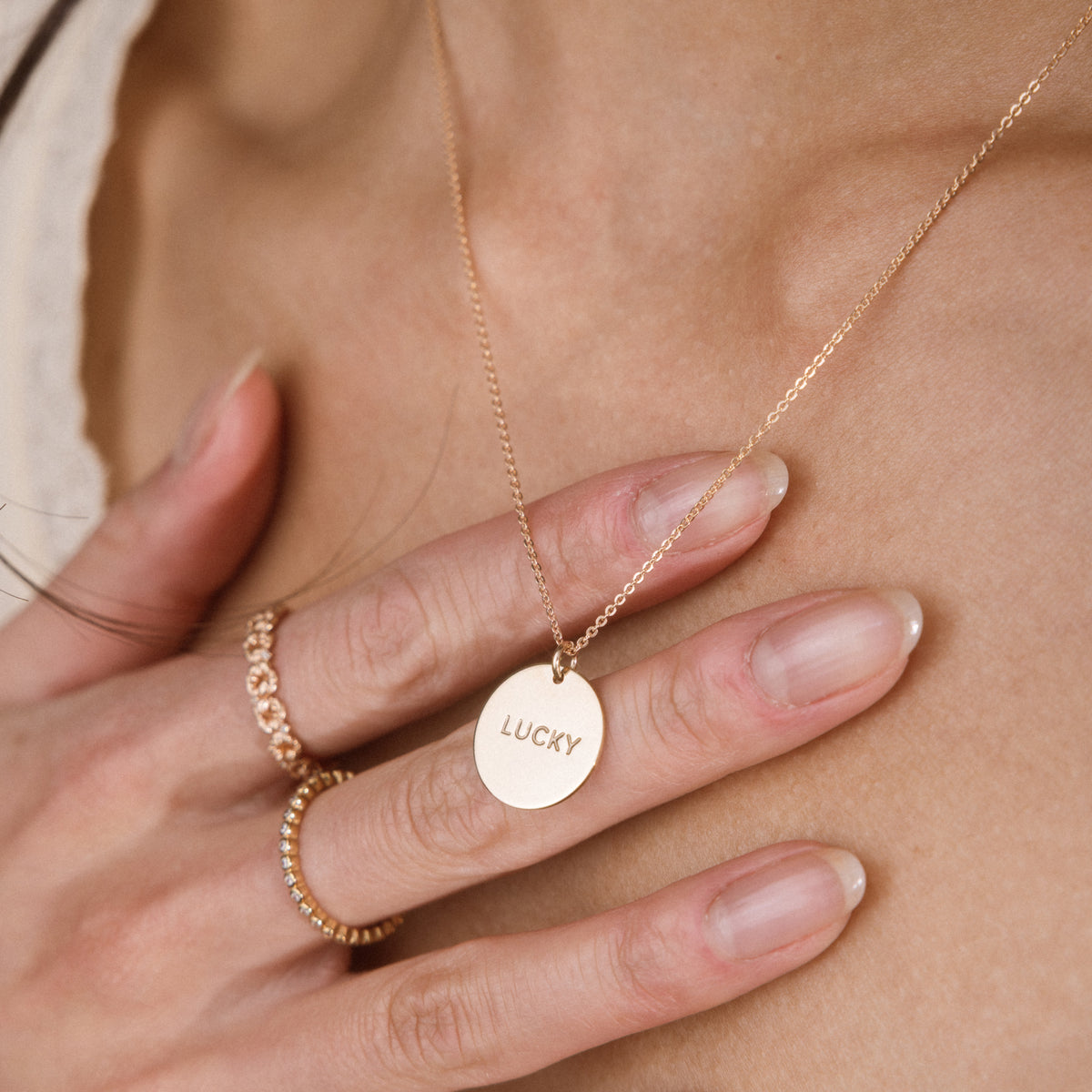 A close-up of a person's hand and neck wearing a delicate gold chain necklace with a round engraved disc pendant that reads 'LUCKY'.