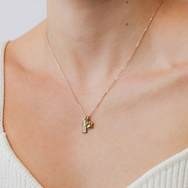 A close-up of a person's neck wearing a gold pendant chain necklace with a small rectangular engraved charm with the name 'ZOE' and a gold polished paw print charm.