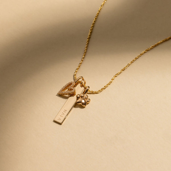 A gold pendant chain necklace with a gold heart charm covered in clear gemstones, a gold thin bar charm with the name 'NALA' engraved, and a gold paw print charm, laying on a solid beige background with shadows.