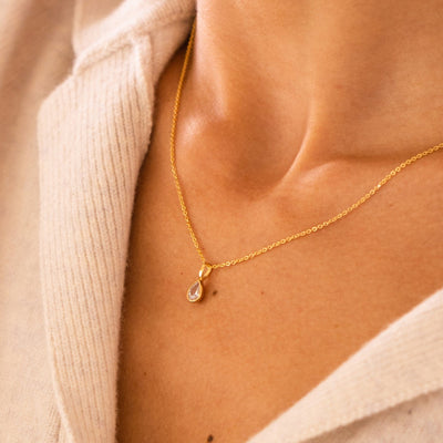 A close-up lifestyle image of a person wearing a delicate gold pendant chain with a pear shaped bezel-set clear gemstone charm and a beige knit collared shirt.