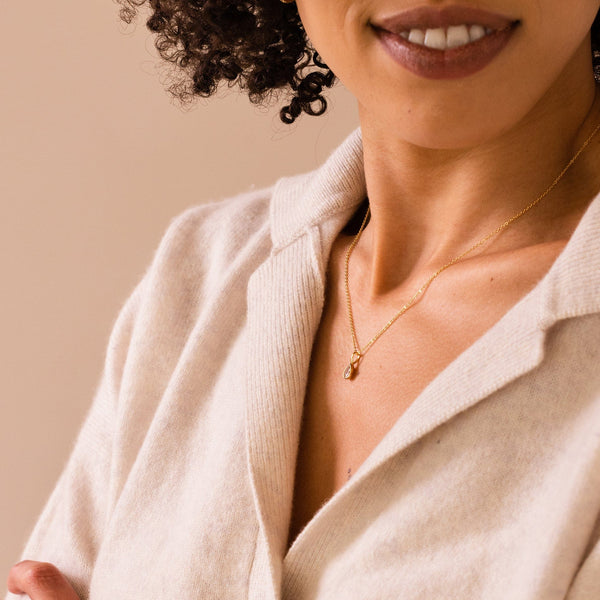 A close-up of a woman in a white knit collared shirt wearing a delicate gold chain necklace with a clear teardrop gemstone charm.