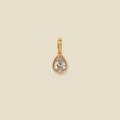 A gold teardrop-shaped pendant with a clear faceted gemstone, centered on a solid beige background.