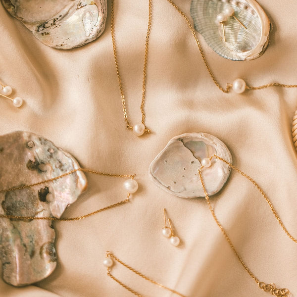 Multiple delicate gold necklaces, bracelets, and earrings laying on a light pink satin surface with oyster shells. Each of the pieces has a single round cream colored pearl.