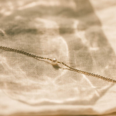 A delicate gold chain bracelet with a single cream colored pearl in the center, laying on a cream colored linen surface.
