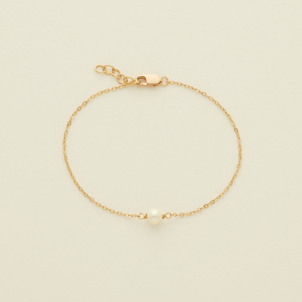 A delicate gold chain bracelet with a single white pearl at the center, displayed on a light cream background.