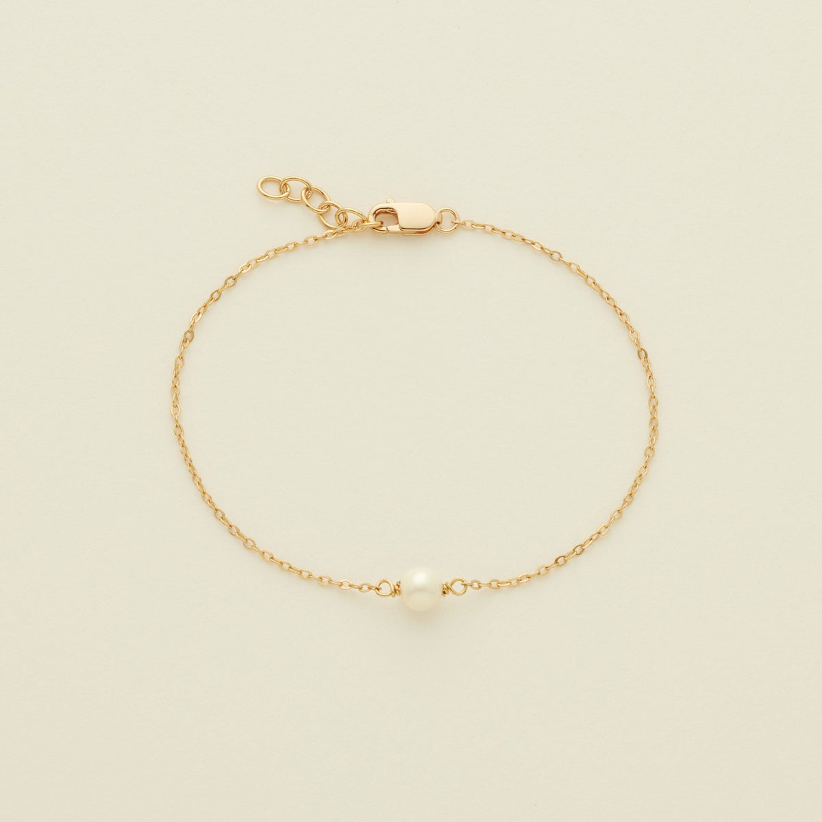 A delicate gold chain bracelet with a single white pearl at the center, displayed on a light cream background.