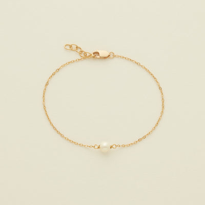 A delicate gold chain bracelet with a single white pearl at the center, displayed on a light cream background.